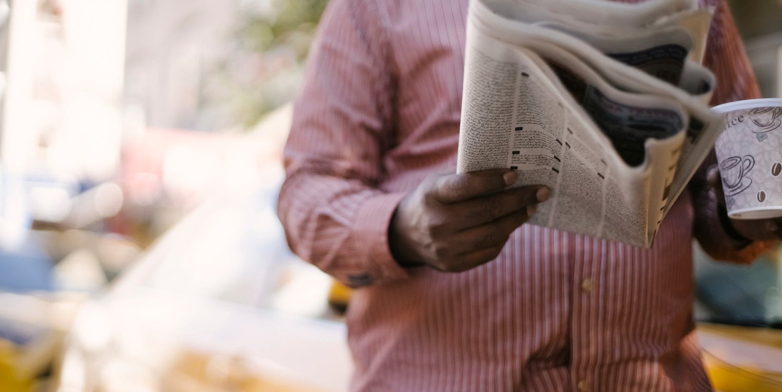 Crop unrecognizable male reading newspaper and holding paper cup with hot beverage while standing on city street