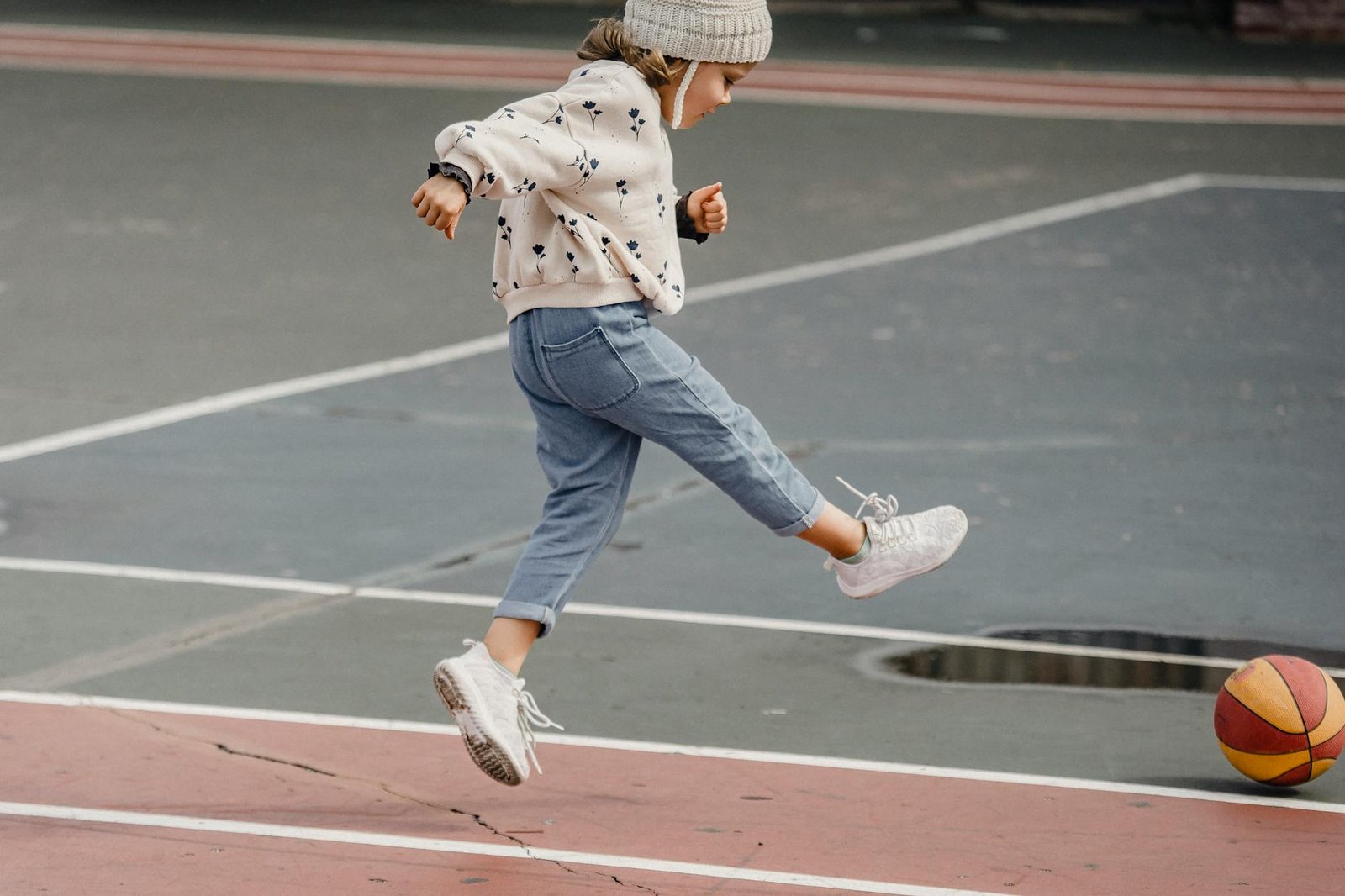 Side view of jumping little girl in jeans and jumper kicking colorful ball playing on sports yard