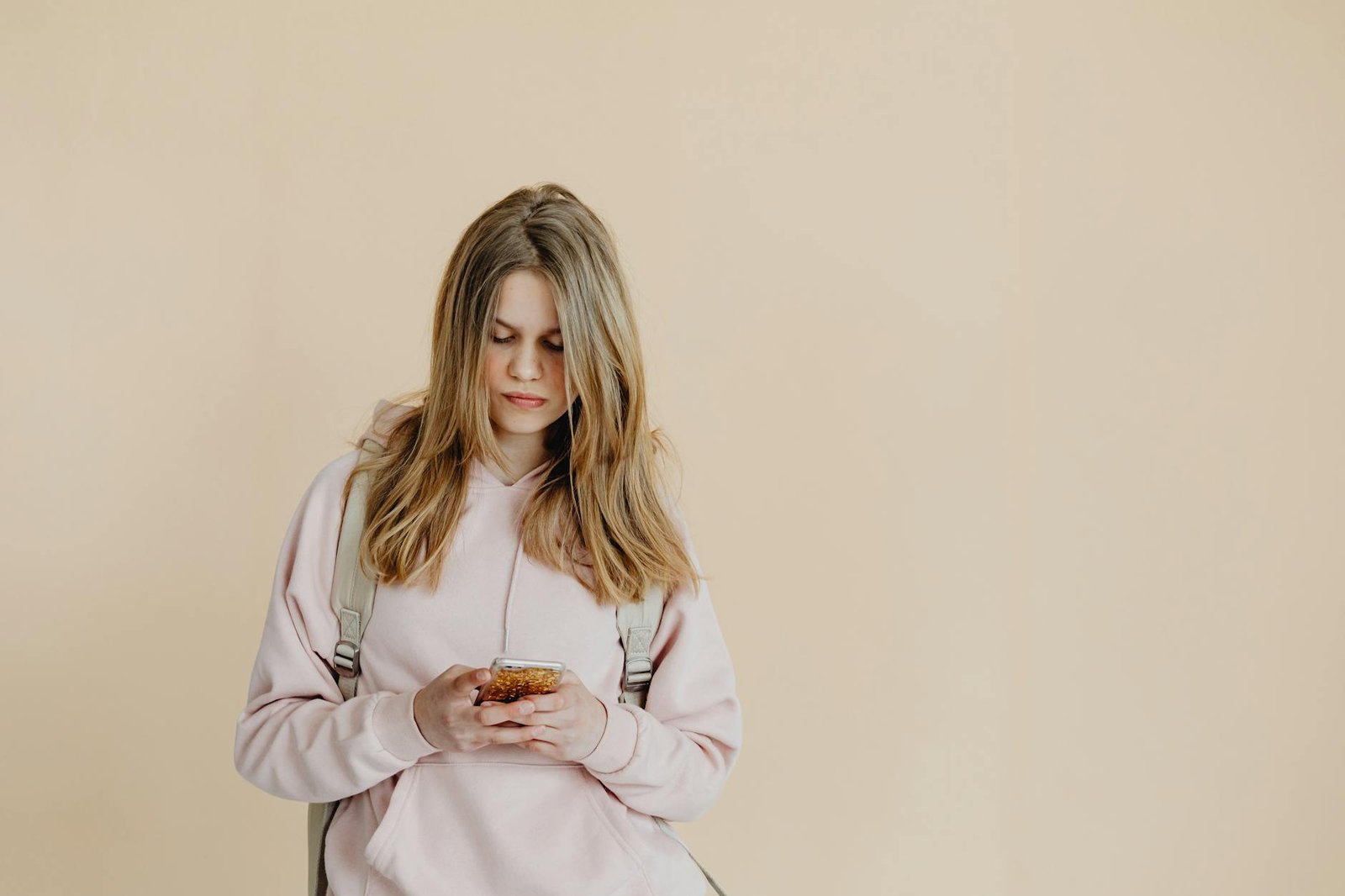 Teenage girl in hoodie and backpack checking smartphone against beige background.